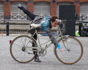 Mary leans across her bike and performs an arabesque, arm pointing towards the floor