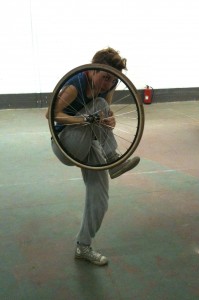 Mary stands on one leg, holding and 'hiding' behind a bicycle wheel