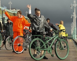 Janine Fletcher & dancers in wet weather gear, stand behind bikes & raise their arms, against a black sky