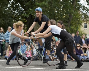 three performers hold onto a fourth's bike as he 'learns to ride'