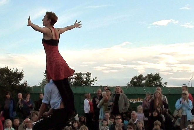 boy cycling around as girl stands on bike saddle with arms outstretched, as if flying