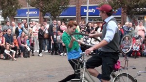 girl spins cycling boy around, both holding onto a bike wheel