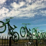 7 green painted bikes leap towards the sky from the top of a fence