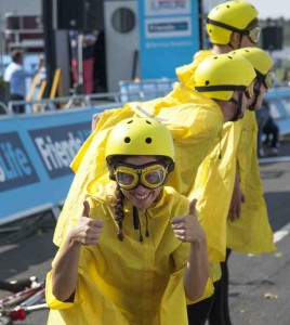 Four performers stand in a line in the Tour de France stage finish line, Bristol. Three performers look away and one smiles to the camera with two thumbs up. Performers wear bright yellow helmets, goggles and cycling capes