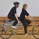 A partially sighted rider balances both feet on the bike frame, whilst the front rider continues to cycle
