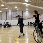 A partially sighted dancer on the ground, pulls a tandem rider by the hand, in a circular loop