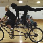 A partially sighted tandem rider stands on the cross bar with one leg raised in an arabesque, whilst the front rider continues to cycle