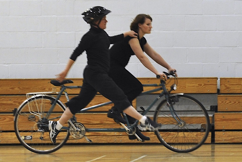 A partially sighted dancer on the ground, leaps alongside a moving tandem, with her hand on the rider's shoulder
