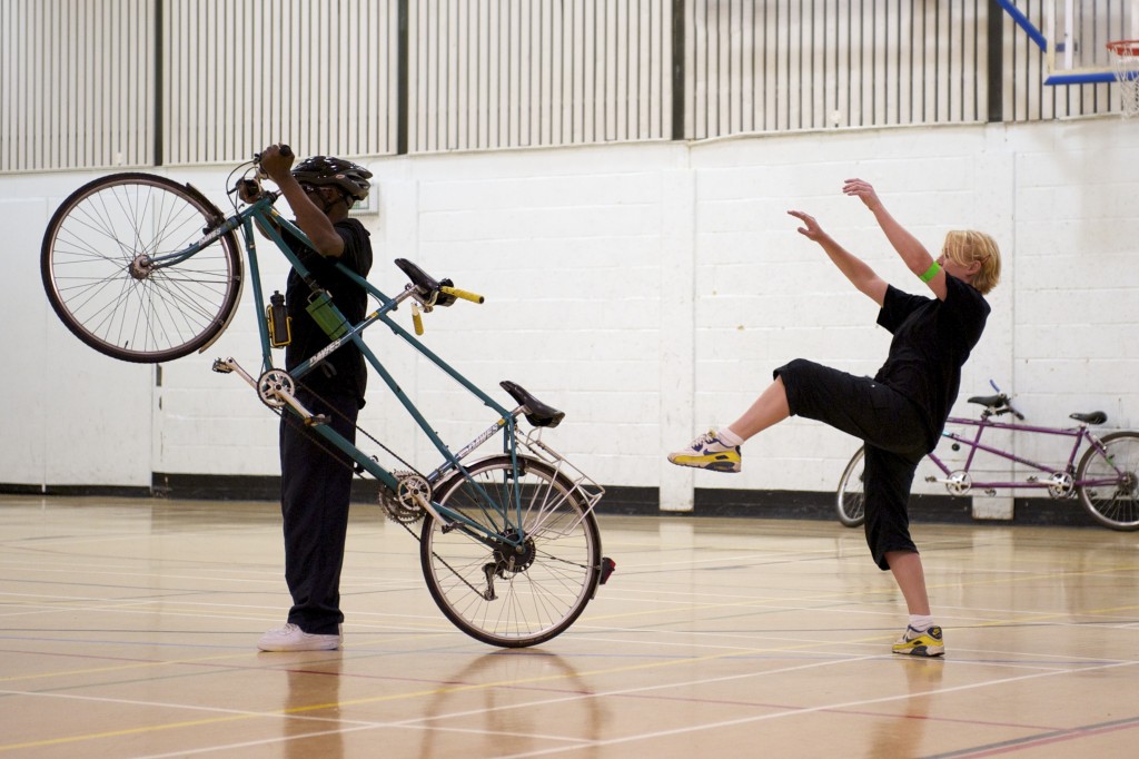 A blind man walks his tandem into the space.  He pulls the bike onto the back wheel and behind, a dancer mirrors, rearing up her leg & arms 
