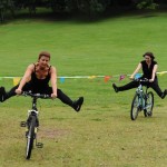 Two performers cycle with legs stretched out to the side