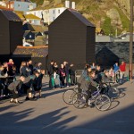 performers on bikes and on the ground, against the backdrop of fishermen's huts and East Hill