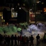 In the performance space, surrounded by audience, performers spin their bikes with green and violet smokes pouring from the back of the bikes