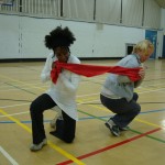 Two participants work together with a scarf, stretching it between them, crouching down and facing away from each other