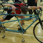 participant holds handlebar and seat of a tandem, with one leg in front he stretches forward