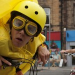 Performer, wearing a yellow helmet, goggles & rain mac, stares into the camera