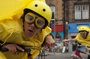 Performer, wearing a yellow helmet, goggles & rain mac, stares into the camera