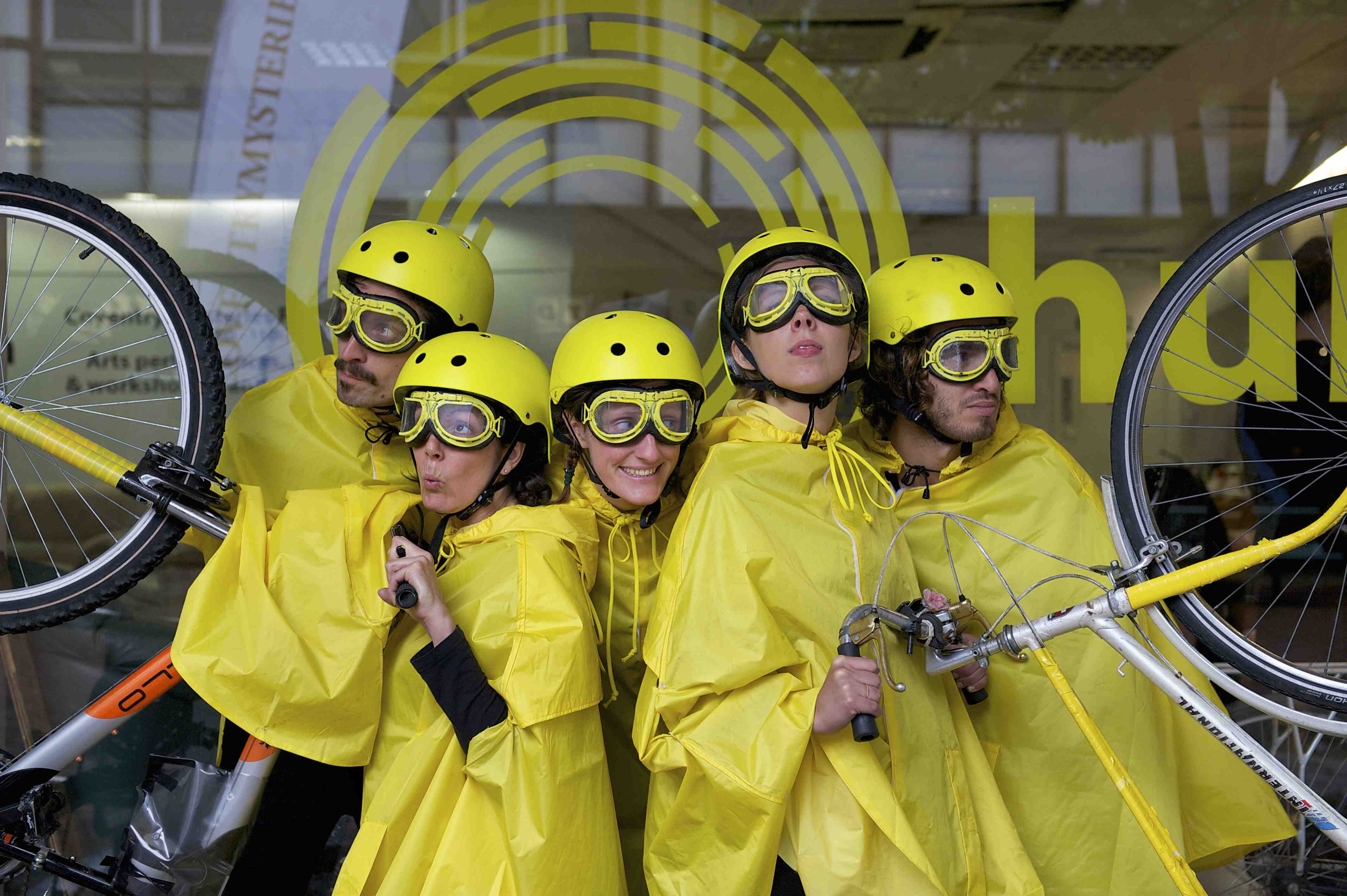 5 performers dressed in yellow cycling rain capes, yellow helmets and yellow goggles, strike a pose with 2 bikes
