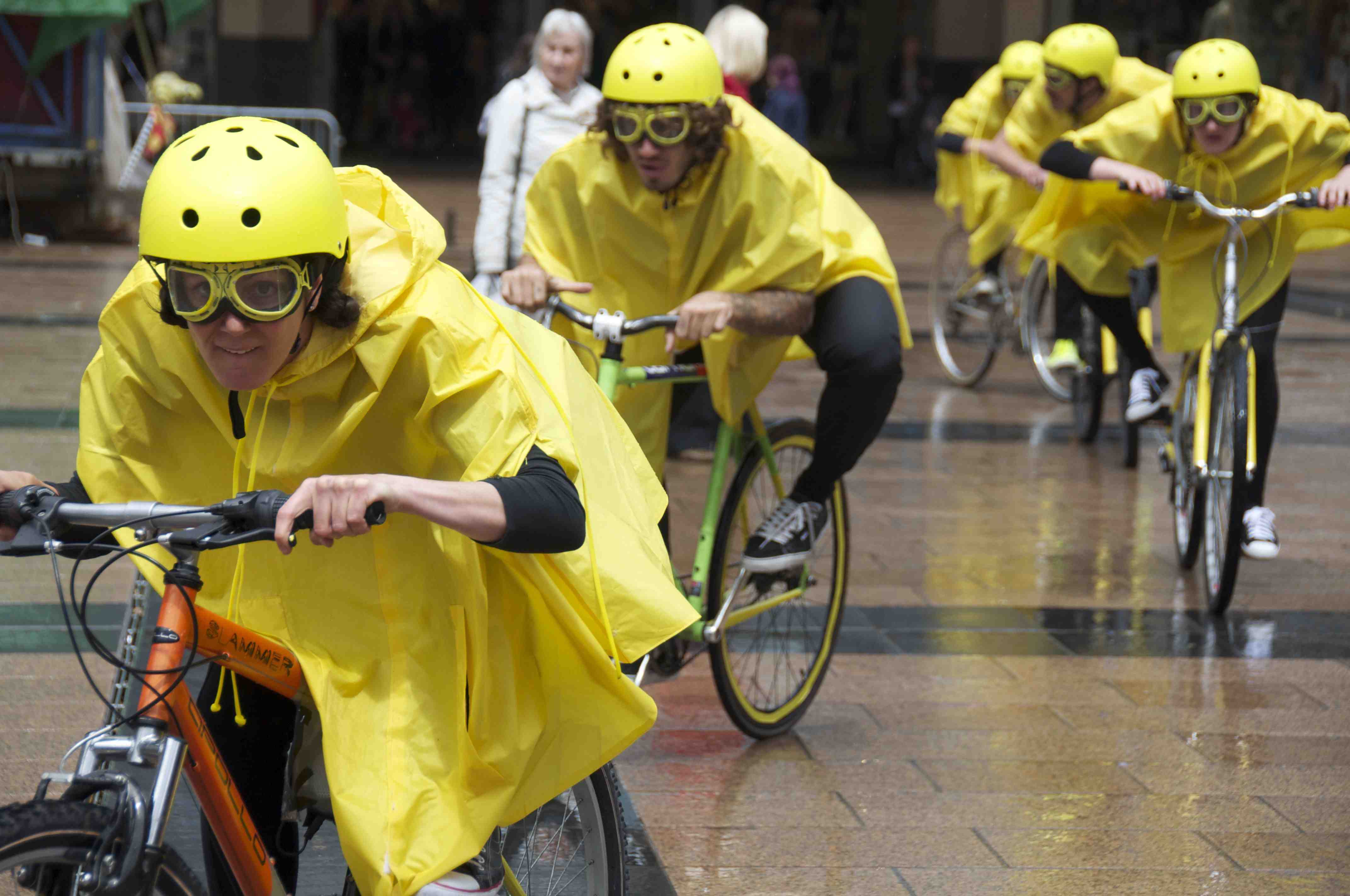 Performers in yellow rain macs, helmets and goggles hunch low over their bike handlebars cycling towards the camera