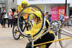 Performer in Yellow helmet, goggles & rain mac kneels behind her upside down bike, peering through the wheel