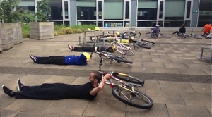 A curving line of workshop participants lie on the floor relaxing, with their heads resting on the saddles of their laid down bikes