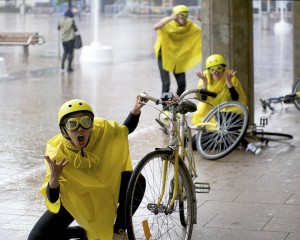 A performer dressed in yellow cape, helmet & goggles kneels on the floor next to her bicycle, berating an imaginary driver who has knocked her off her bike!